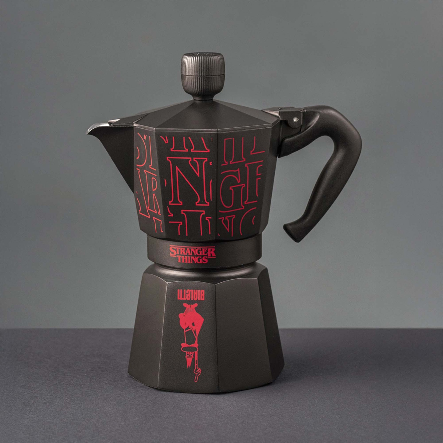 Una caffettiera moka nera con il logo rosso di Stranger Things. Ha un manico e un coperchio. Lo sfondo è grigio.