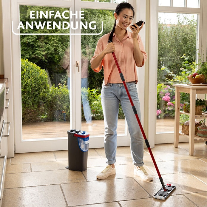 Eine Frau wischt einen Steinboden. Sie hält einen roten Wischmopp. Ein Eimer und ein Fenster sind hinter ihr. Sie telefoniert.