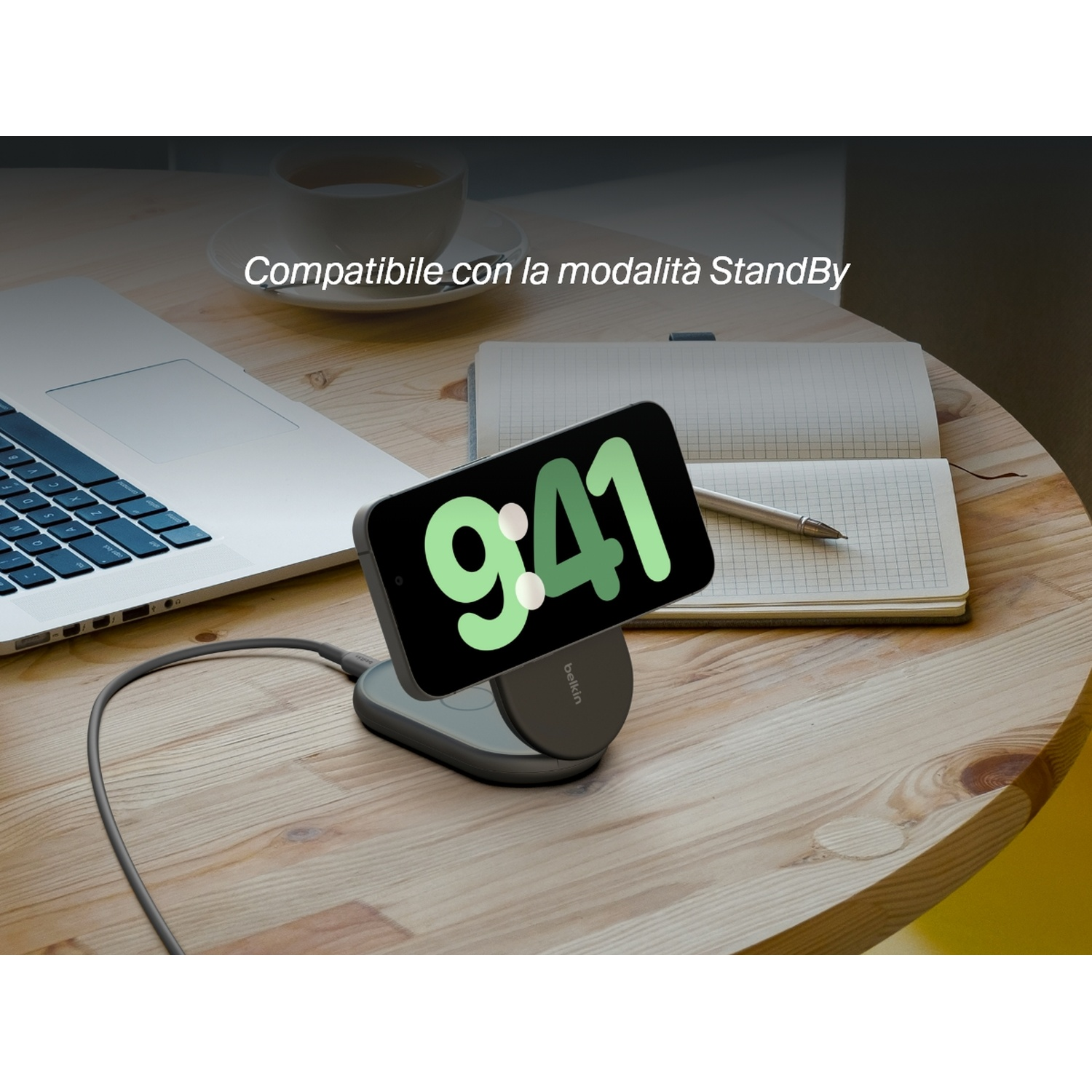 Un iPhone che mostra 9:41 su un caricatore wireless, con un laptop, un quaderno e una penna su un tavolo di legno. Il testo dice 'StandBy'.