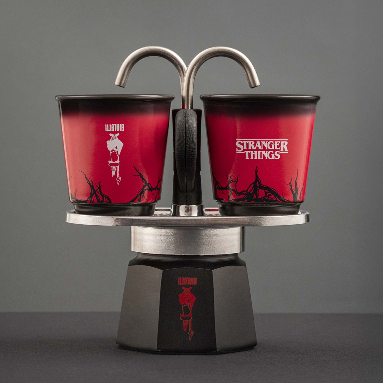 Una caffettiera espresso Stranger Things rossa e nera con due tazze. Le tazze hanno il logo Stranger Things e il logo Bialetti.