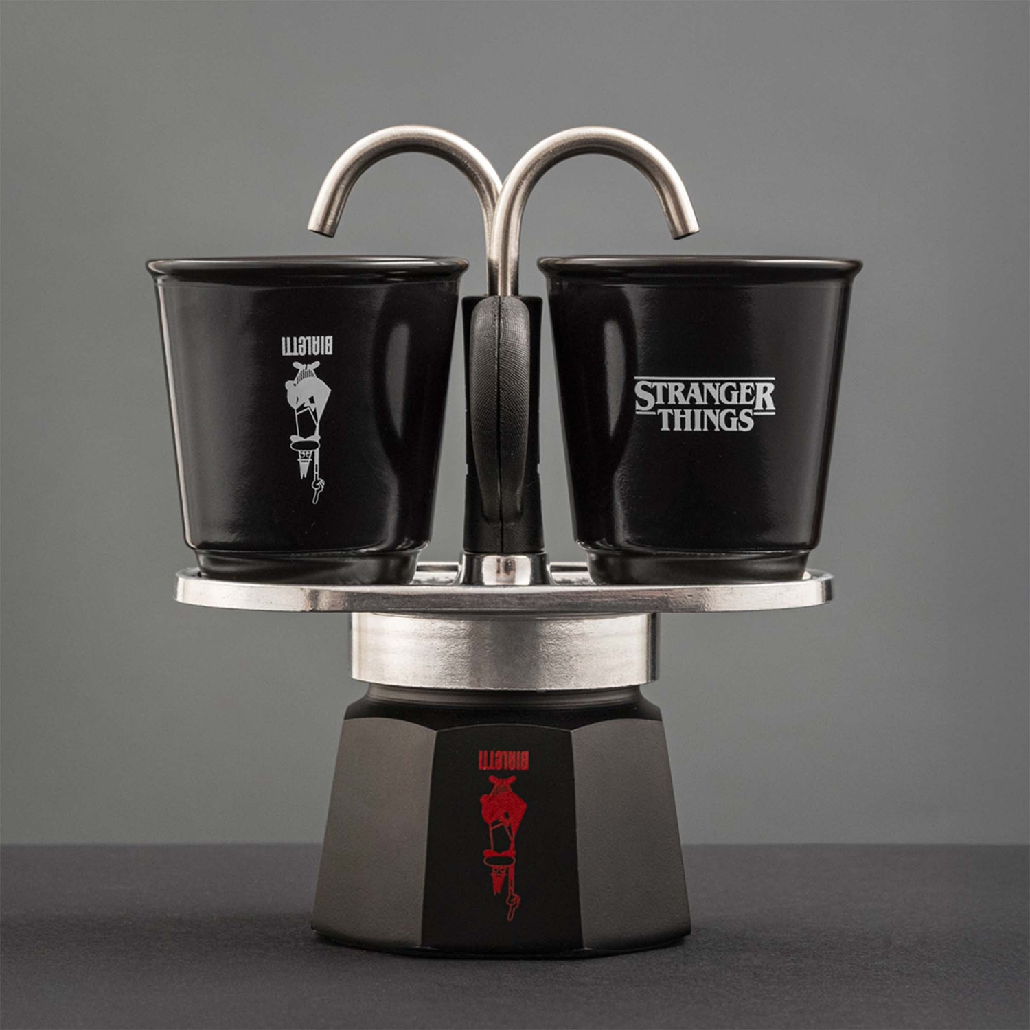 Caffettiera espresso nera con due tazze. Una dice 'Bialetti', l'altra 'Stranger Things'. Dettagli e manico argentati, sfondo grigio.