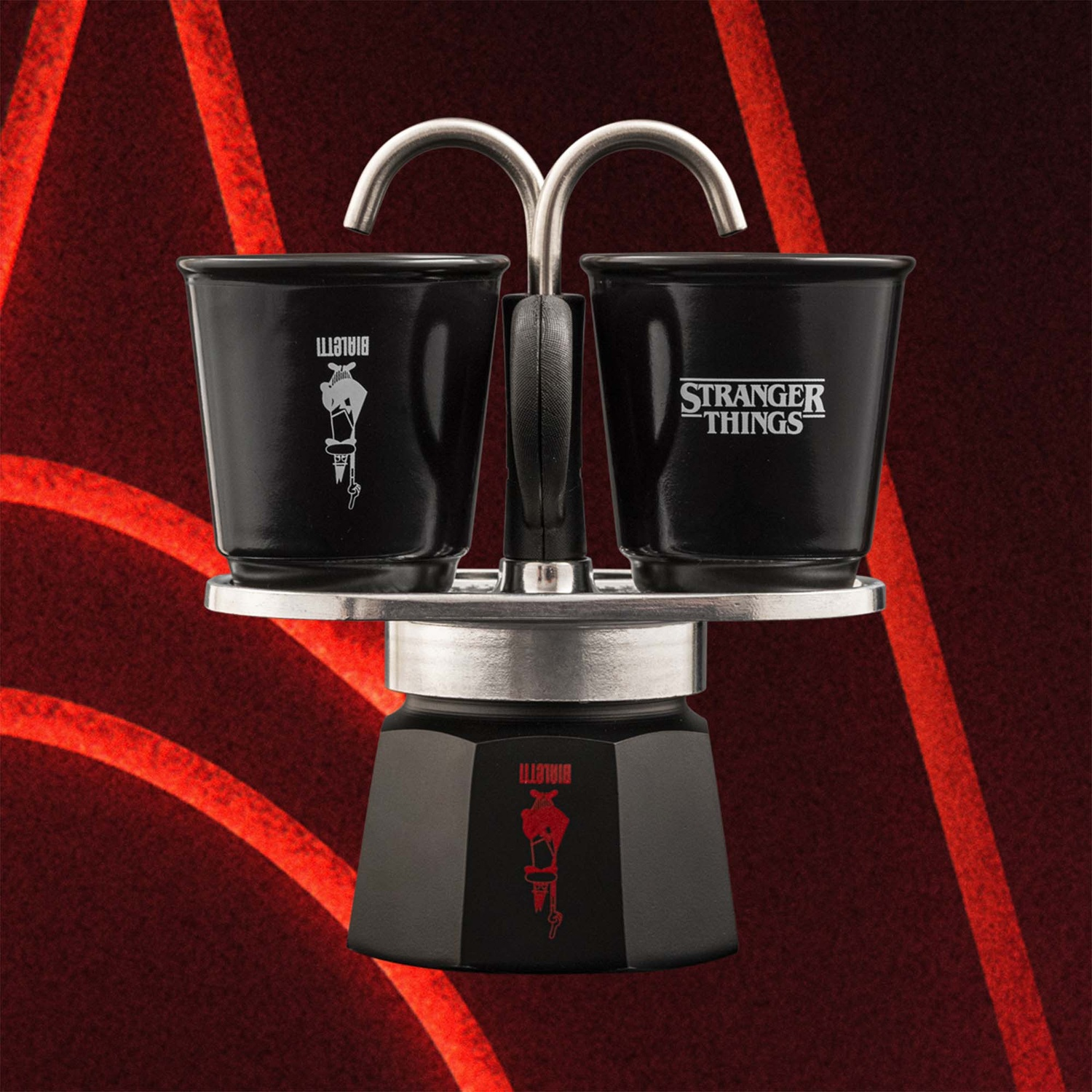 Macchina per caffè espresso nera con due tazze, una con 'Bialetti', l'altra 'Stranger Things'. Sfondo rosso con linee curve.