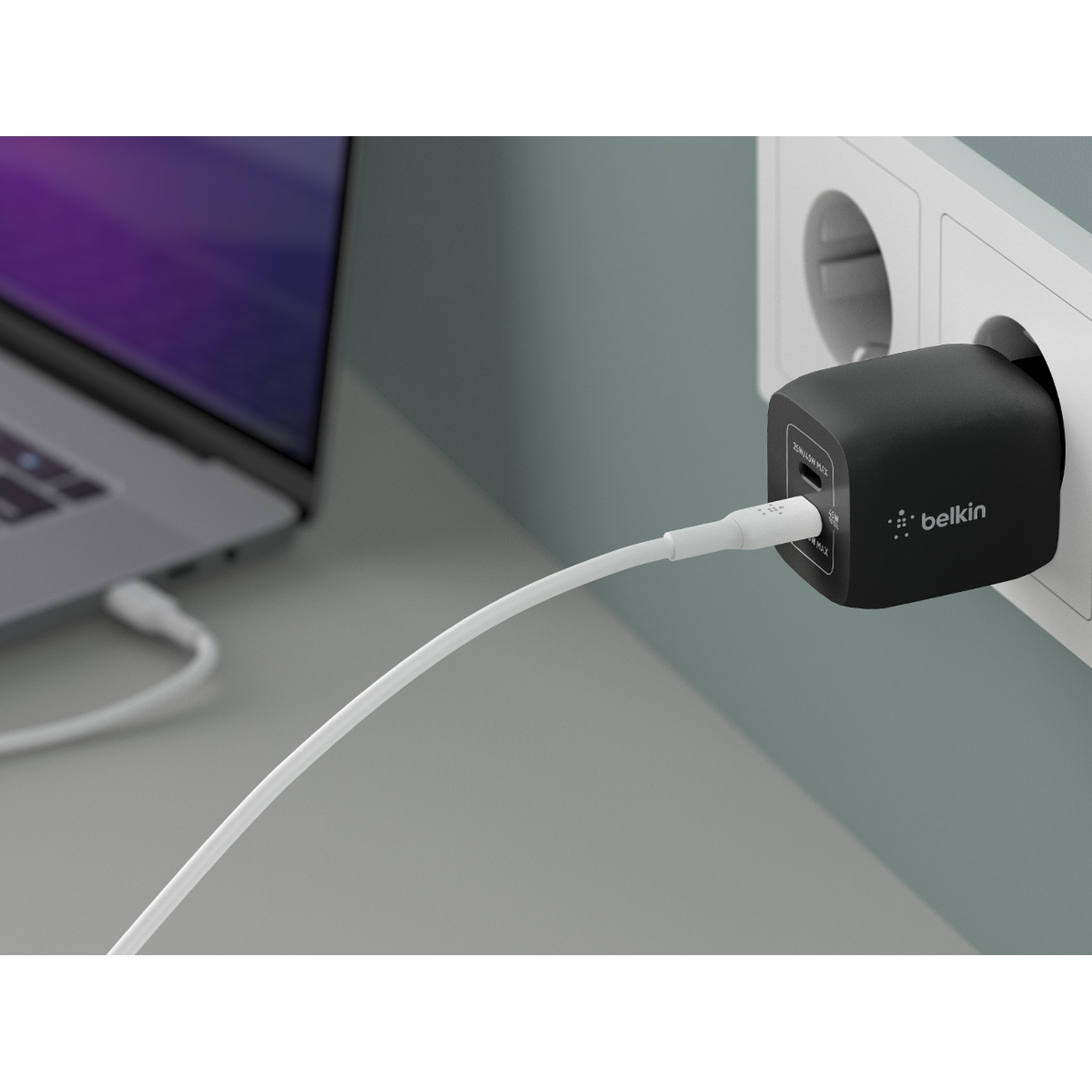 Un caricabatterie Belkin nero collegato a una presa a muro. Un cavo bianco collega il caricabatterie a un laptop a sinistra.