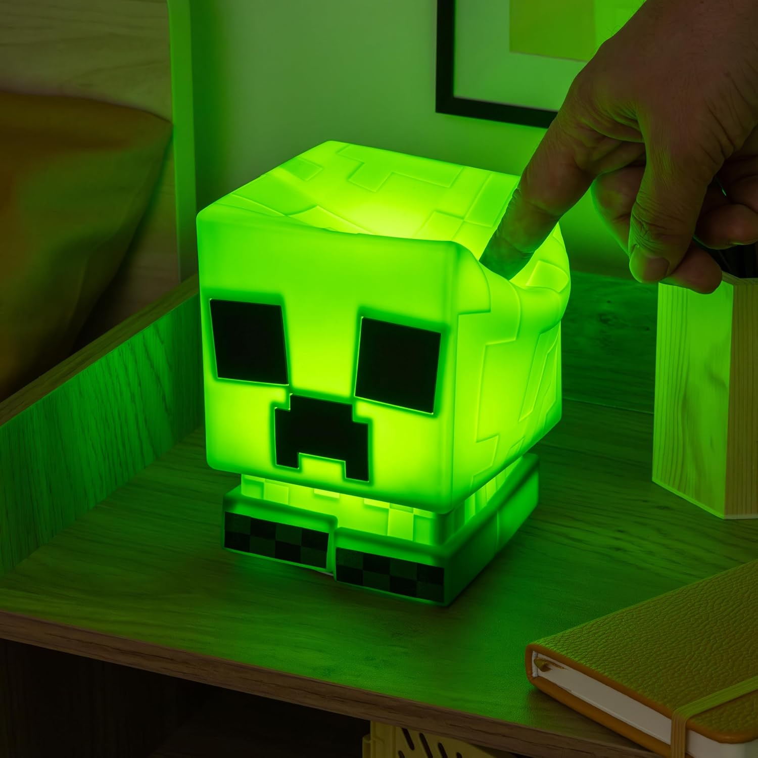 Egy zöld Minecraft Creeper lámpa egy fa felületen. Egy kéz megérinti a lámpát. Közelben egy könyv és egy fatömb.