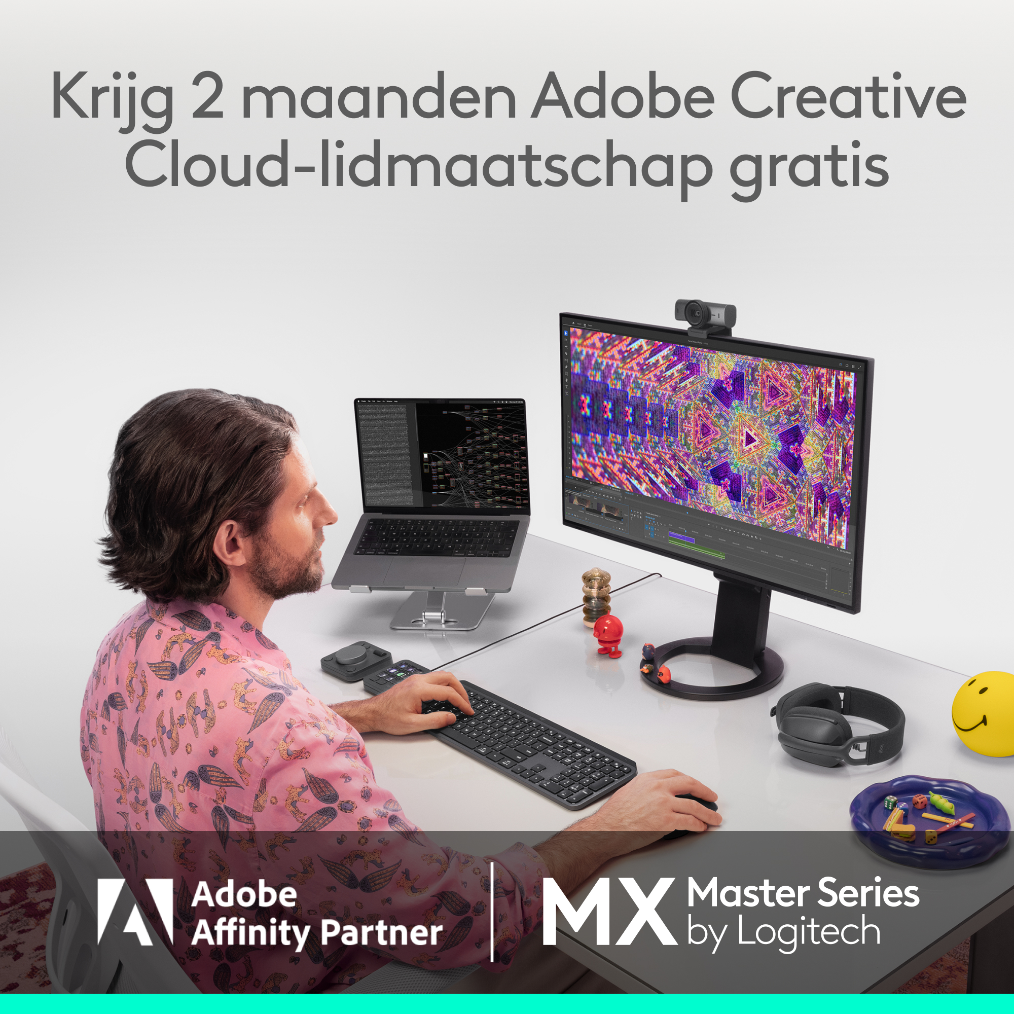 Man at desk uses computer. Monitor shows colorful graphics. Laptop, keyboard, headphones, and other items on the desk.