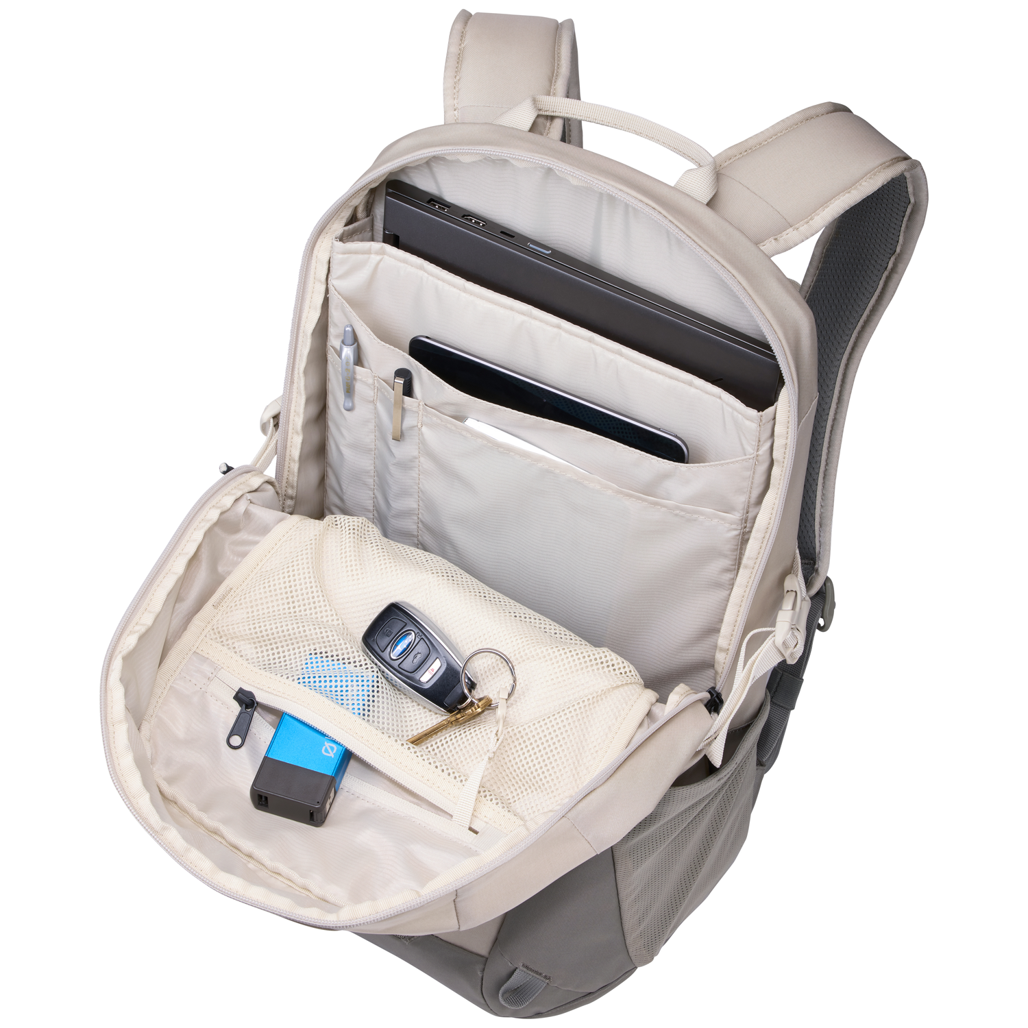 Open backpack with laptop, tablet, pens, keys, and charger visible. Beige and gray colors dominate the design, with mesh pockets.