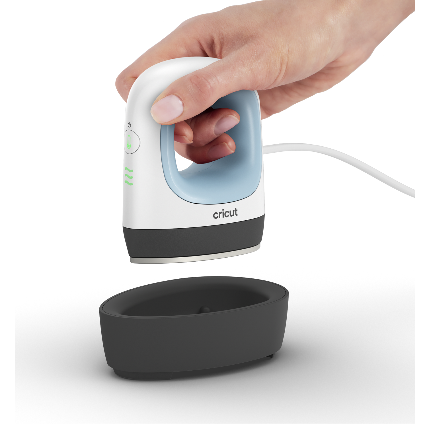 A hand holding a white and blue Cricut iron above a black base. The iron is plugged in, ready to use.