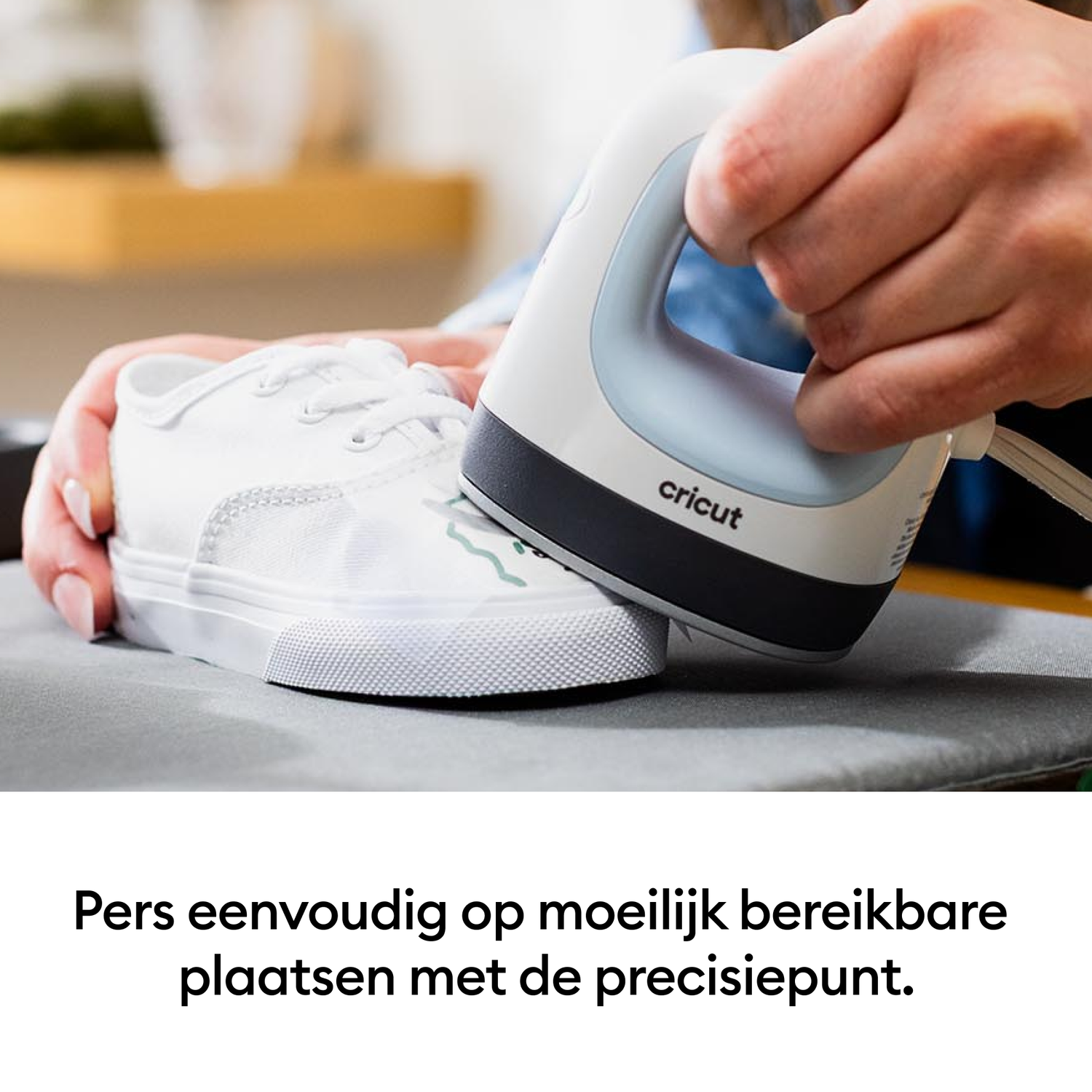 A person uses a Cricut heat press on a white shoe. The press is held over a design on the shoe. The shoe is on a gray surface.