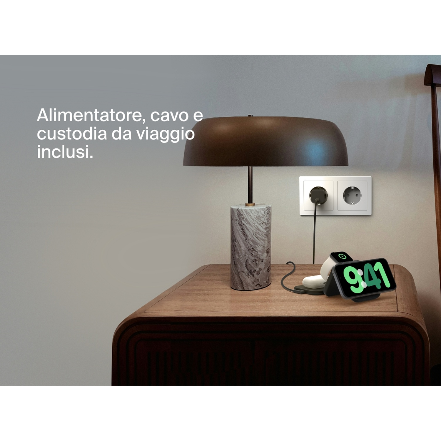 Una lampada, telefono con display 9:41, caricabatterie wireless e presa a muro su comodino in legno. Telaio letto visibile.