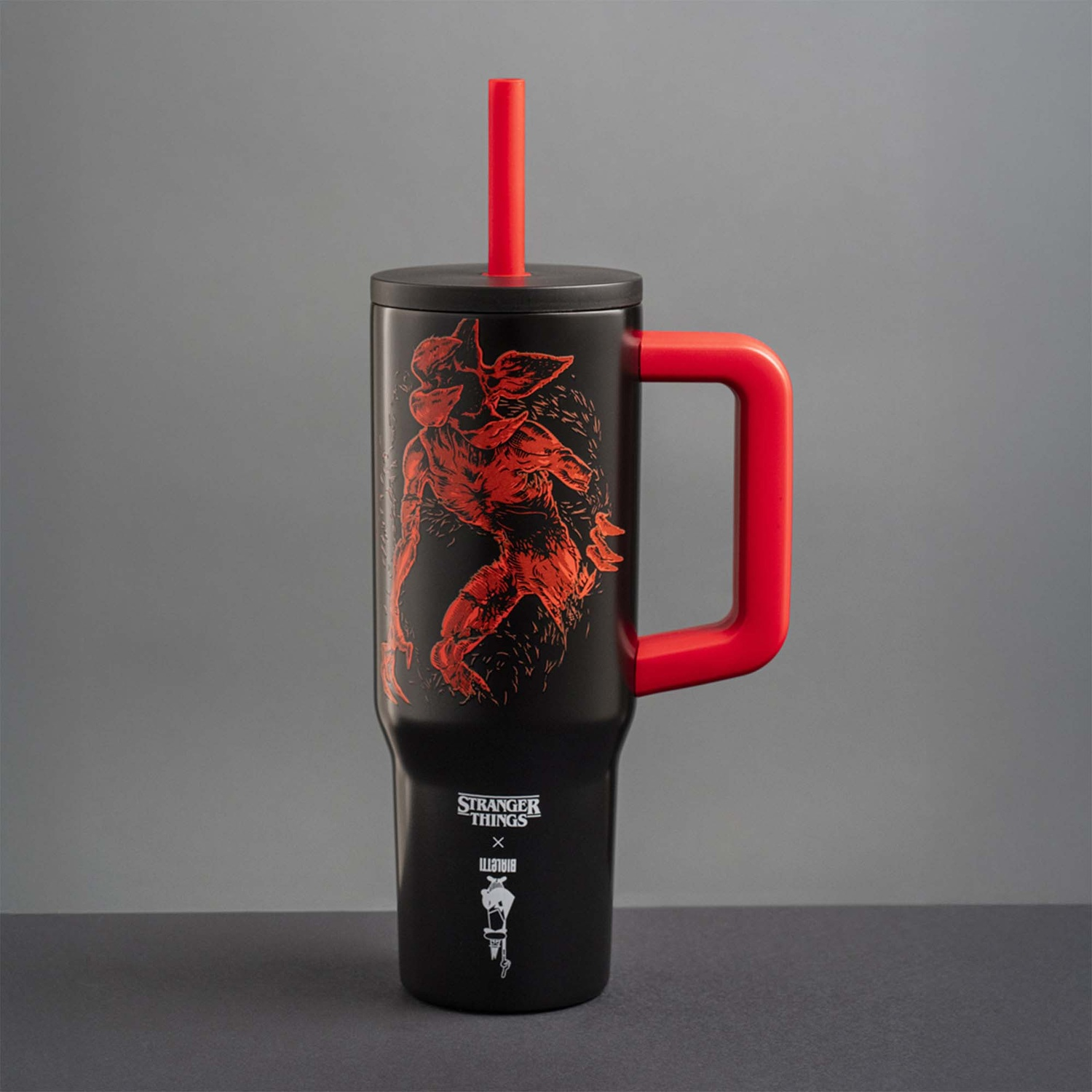 Bicchiere nero 'Stranger Things' con manico e cannuccia rossi. Grafica Demogorgon e logo dello show sul davanti. Sfondo grigio.