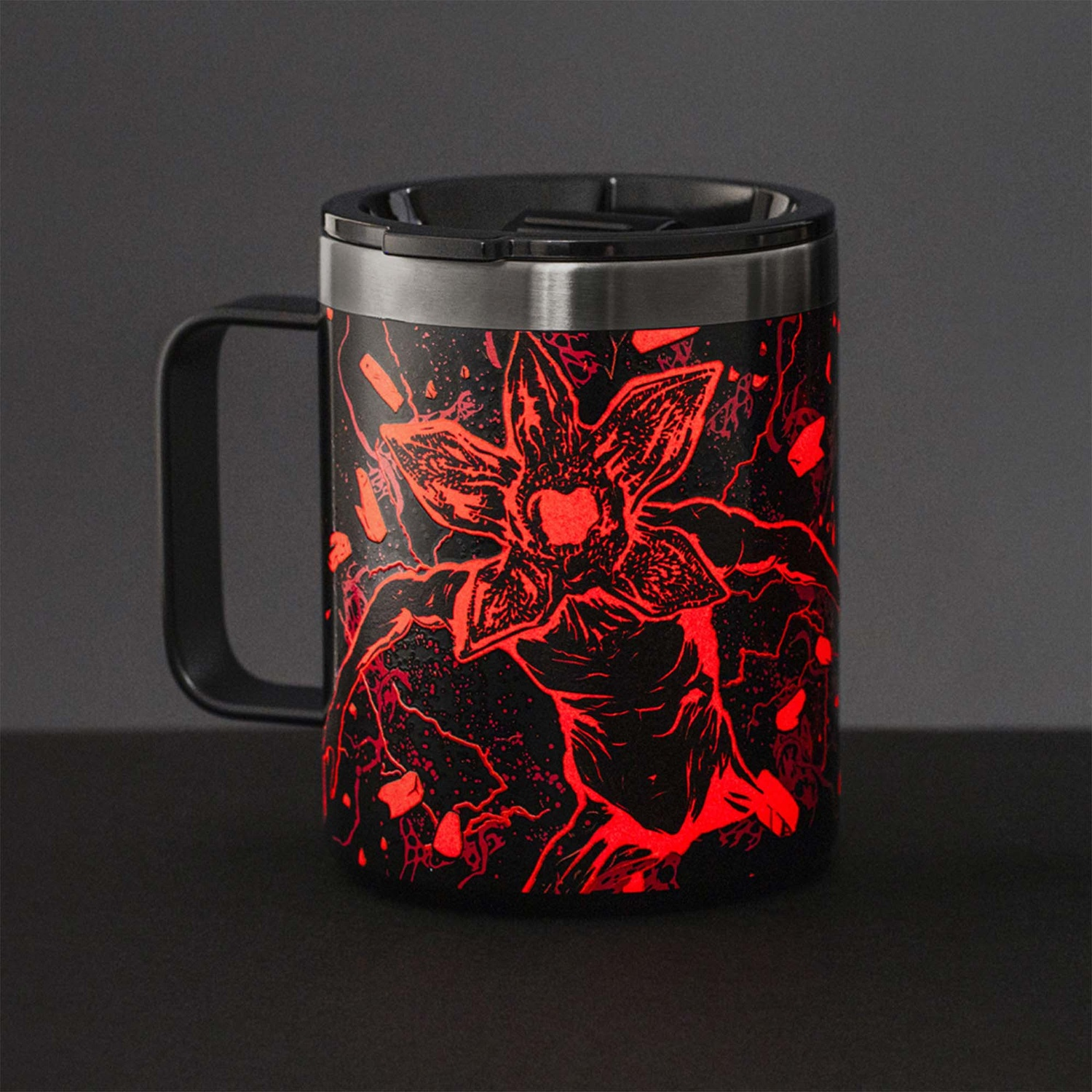 Una tazza da viaggio nera con un disegno rosso Demogorgon. La tazza ha un manico e un coperchio neri. È appoggiata su una superficie nera.