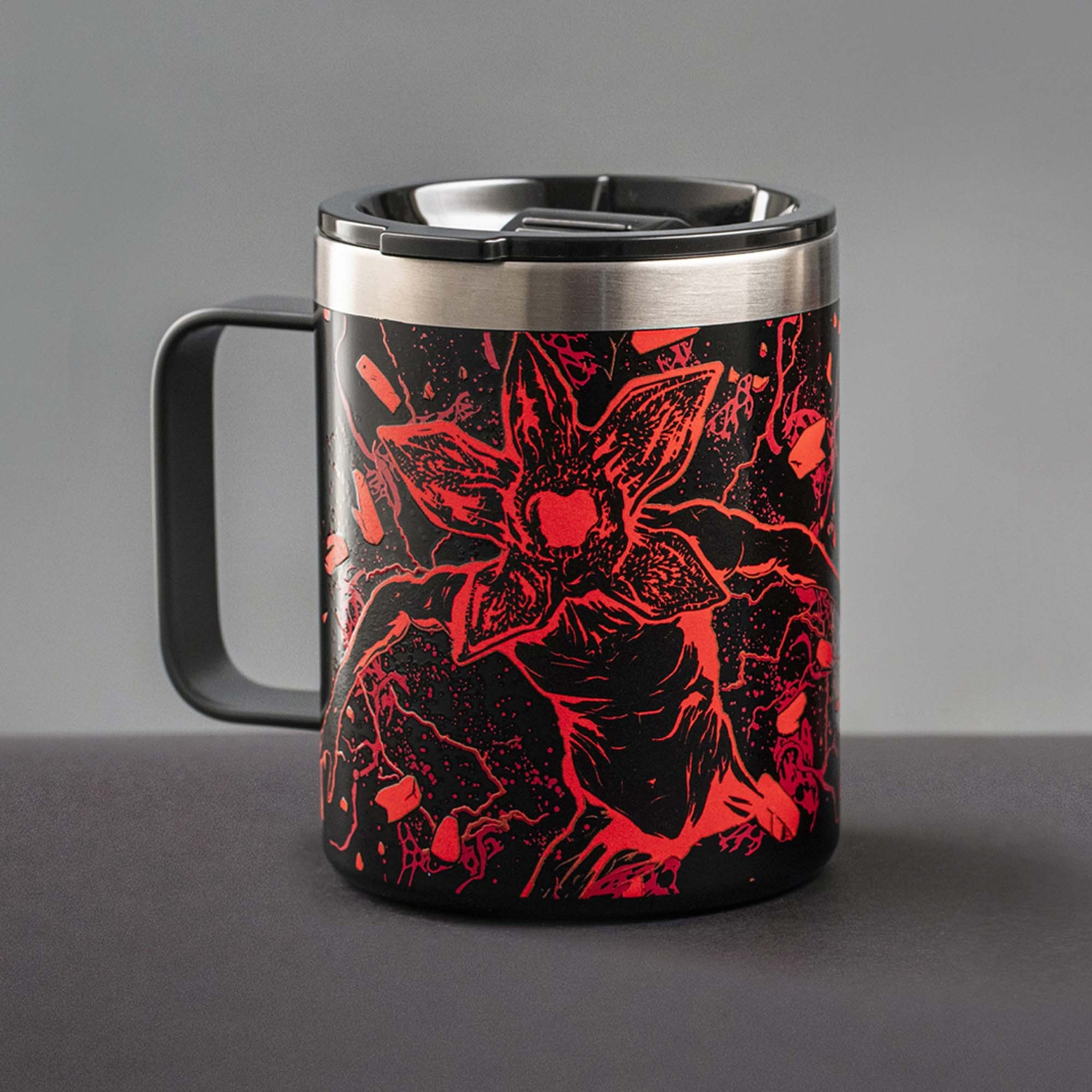 Tazza da viaggio nera con design Demogorgon rosso. Bordo argentato, manico e coperchio neri. Su una superficie scura contro uno sfondo grigio.
