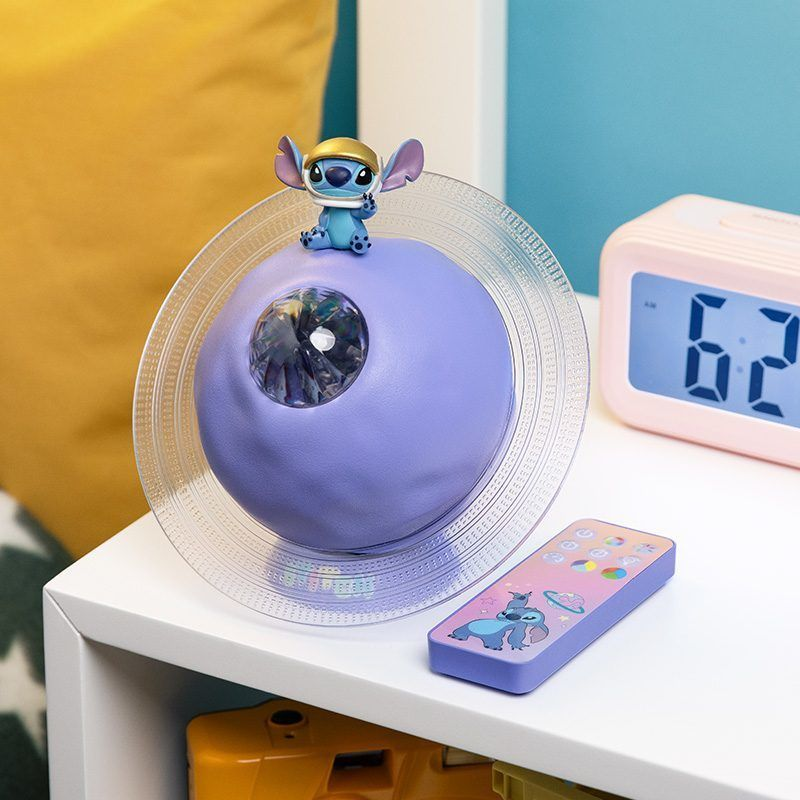 Egy lila Stitch témájú éjszakai fény figura a tetején. Egy távirányító és egy rózsaszín digitális óra is látható.