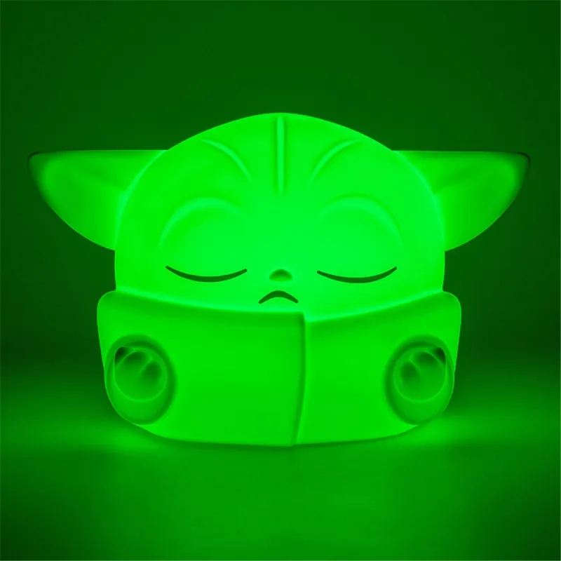 Egy zöld Baby Yoda lámpa világít a sötétben. Csukott szemei és nagy fülei vannak. A háttér egy zöld fény gradiens.