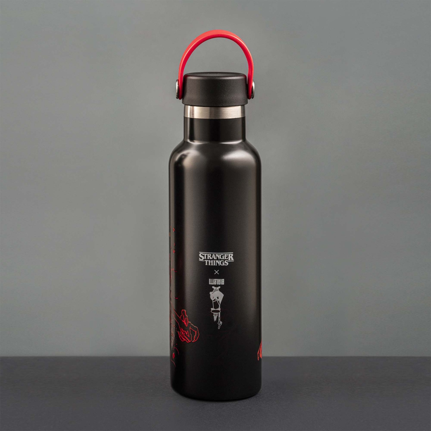 Borraccia nera con manico rosso. Il logo "Stranger Things" e la grafica rossa sono stampati sulla bottiglia. È su una superficie scura.