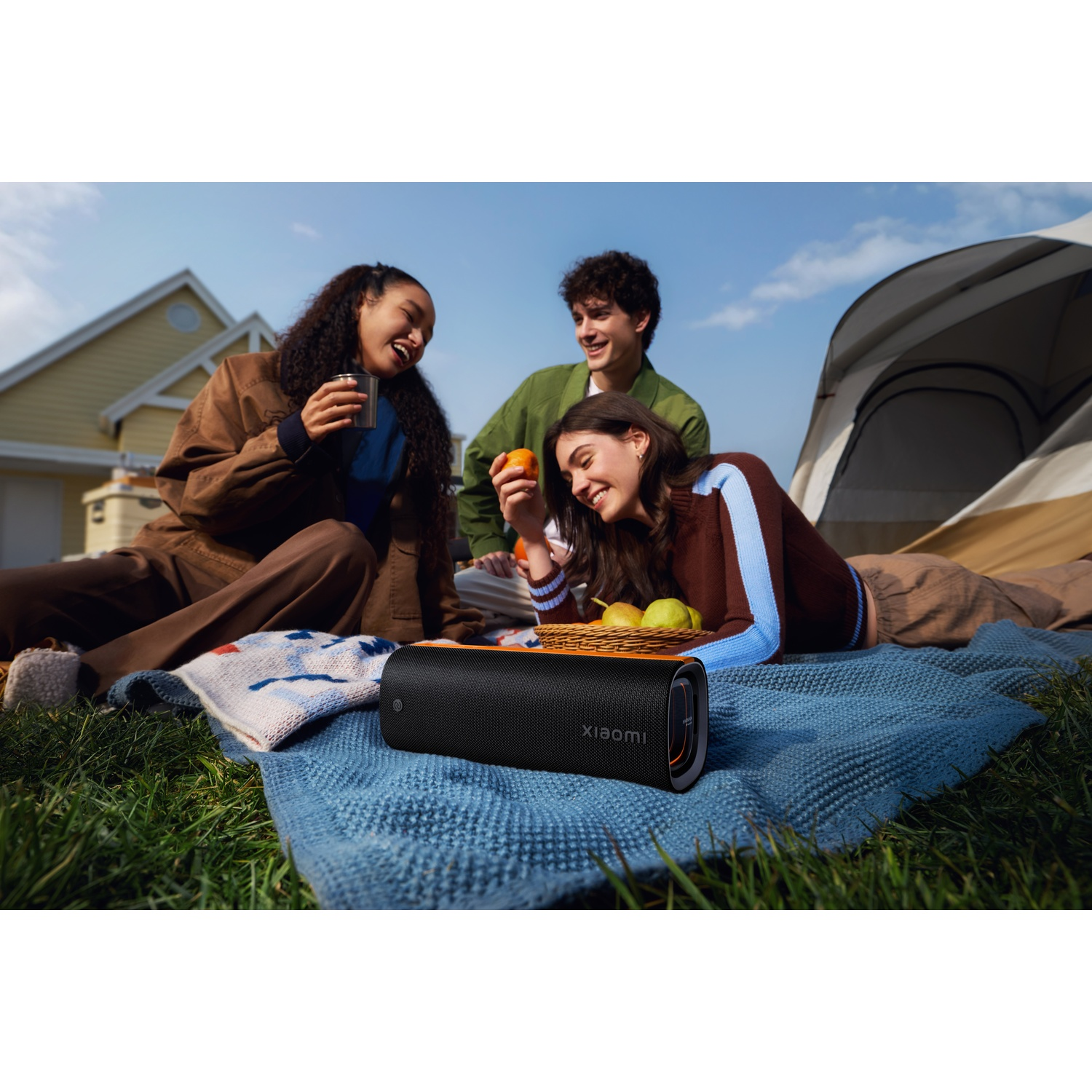 Drie jonge mensen lachen buiten. Eén houdt een kopje vast, een ander een sinaasappel. Picknickdeken, luidspreker, tent en huis op de achtergrond.