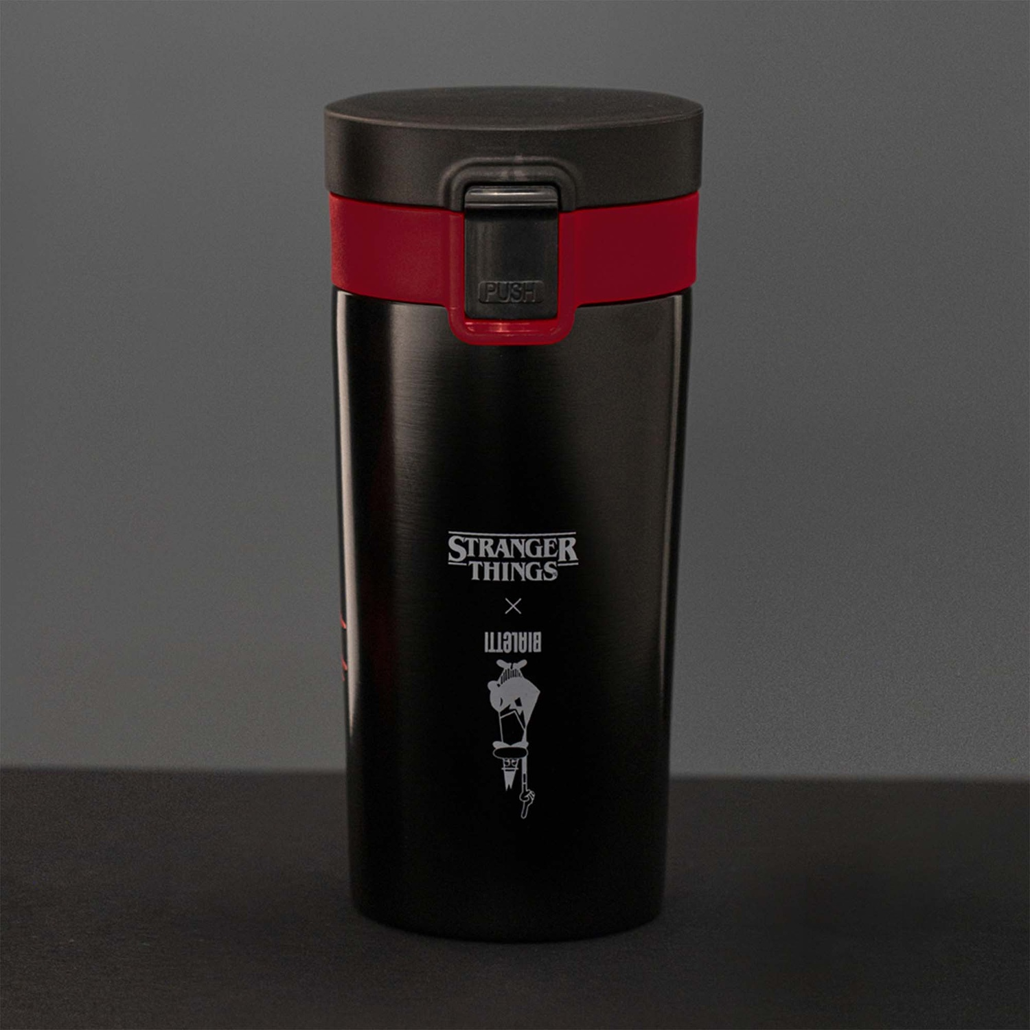 Tazza da viaggio nera di Stranger Things con una fascia rossa. La tazza ha un coperchio nero e un logo di Stranger Things x Bialetti.