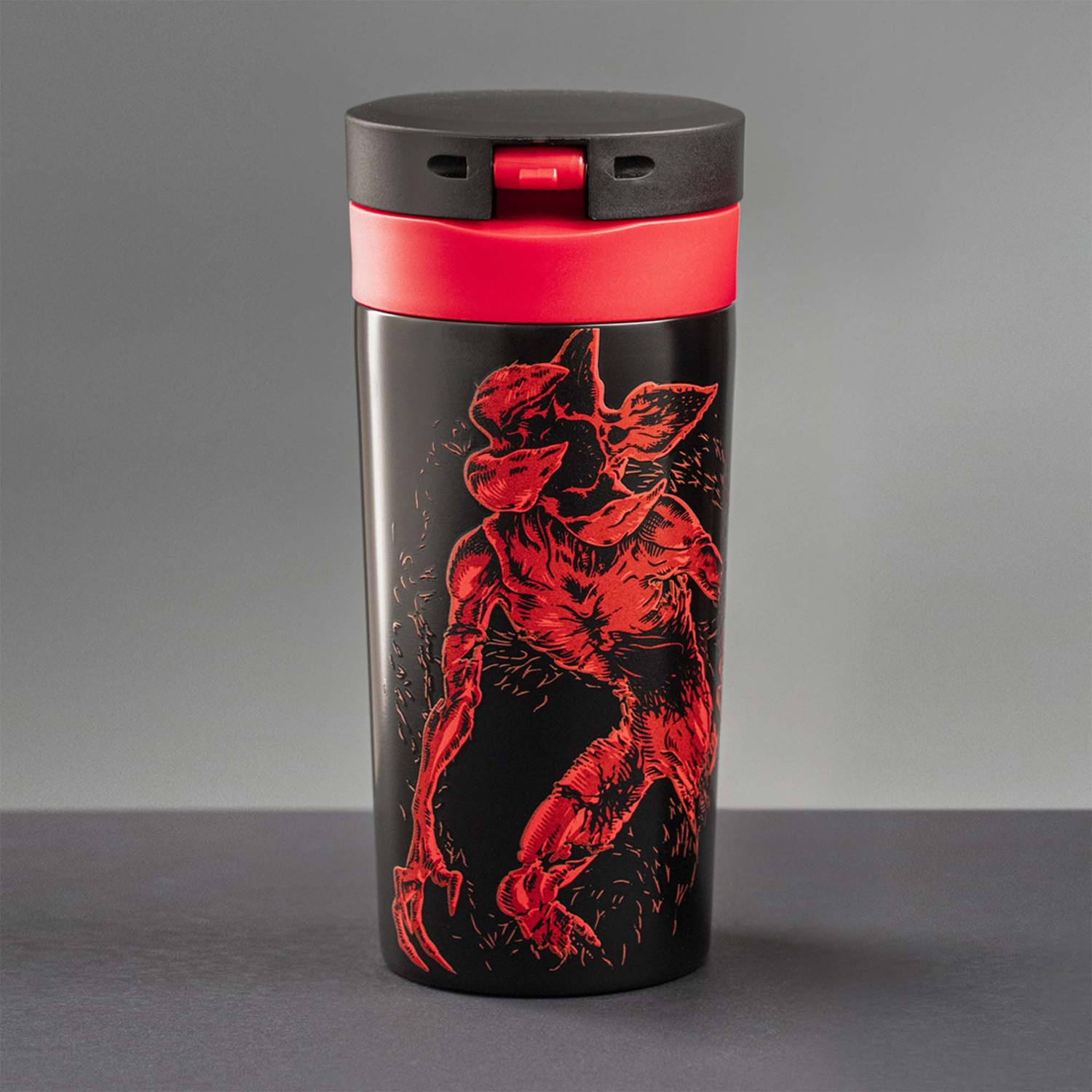 Tazza da viaggio nera con illustrazione rossa di Demogorgon. Ha un coperchio nero e una fascia rossa. Lo sfondo è grigio.