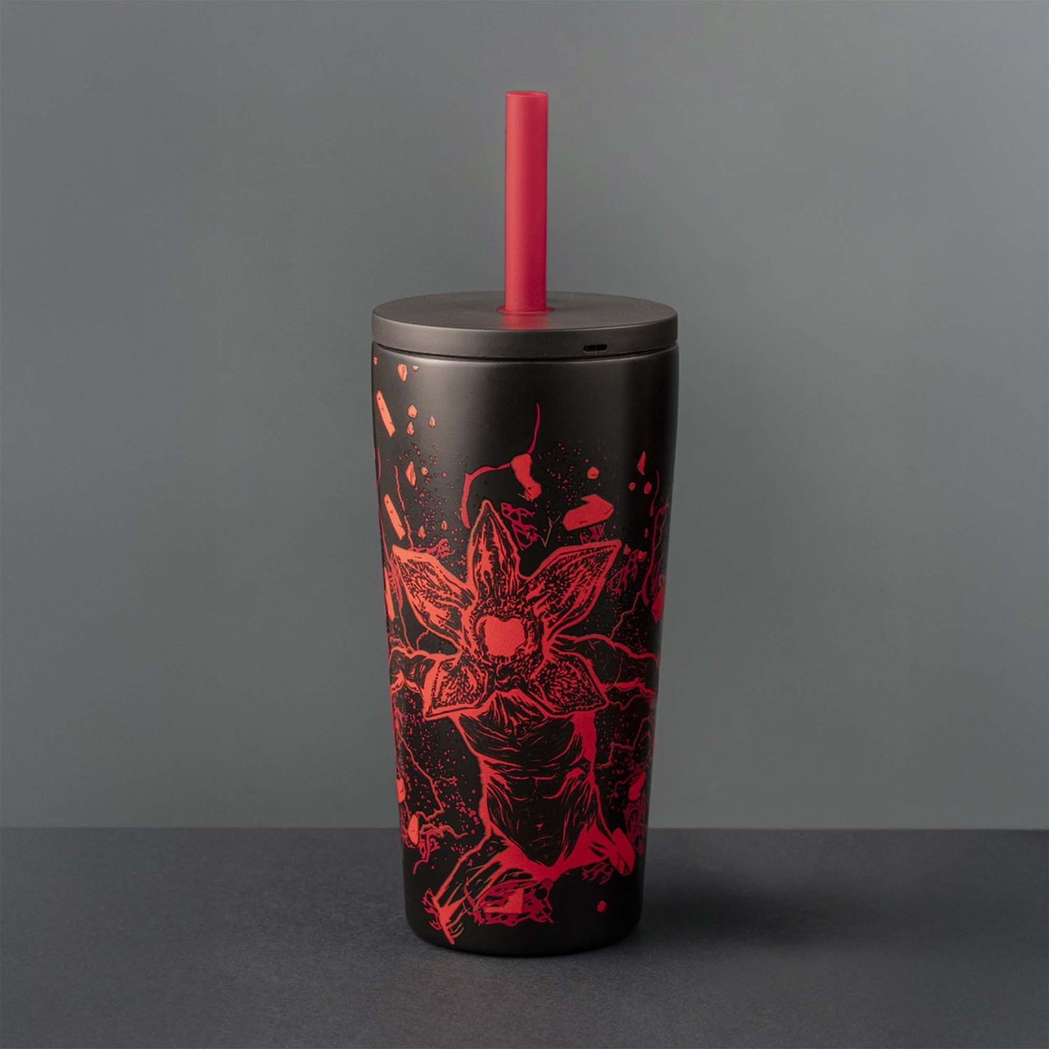 Bicchiere nero con design Demogorgon rosso e cannuccia. È su una superficie scura contro uno sfondo grigio.