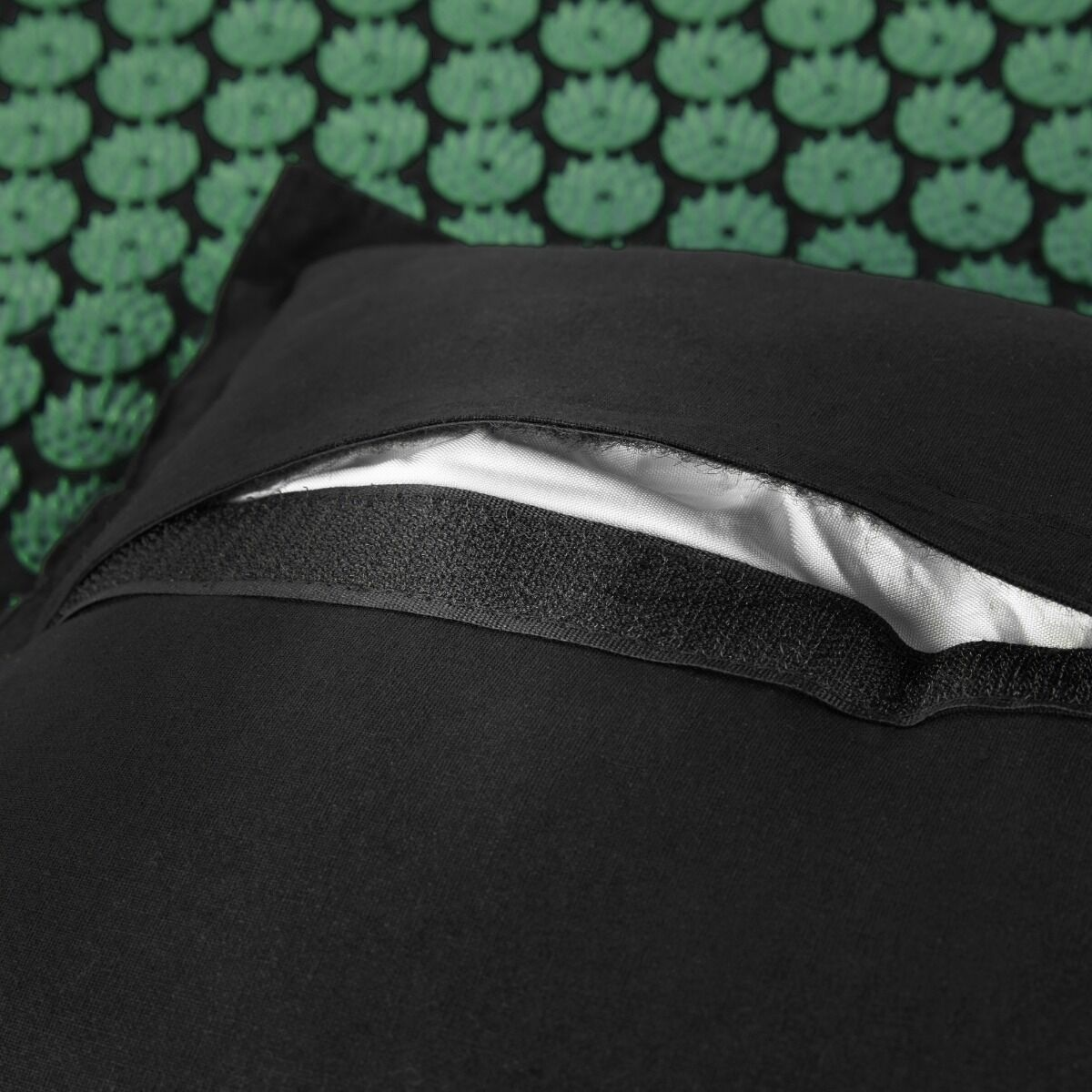 Czarna poduszka z białym wnętrzem, zapięciem na rzep, na tle zielono-czarnego wzoru kwiatowego.