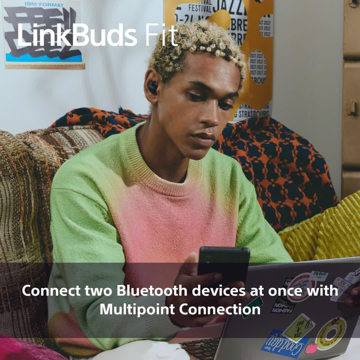 Una persona con auricolari e telefono usa un laptop. La persona indossa un maglione verde e rosa. Un poster è sullo sfondo.
