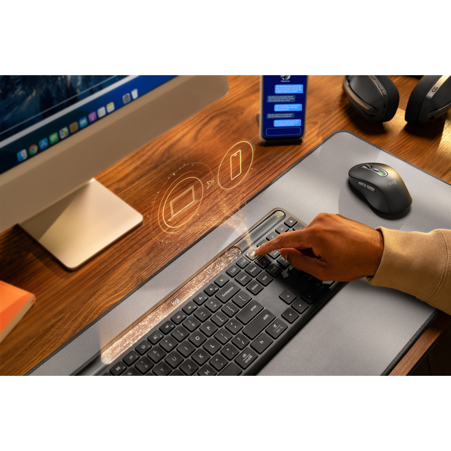 Una mano preme un pulsante su una tastiera. Accanto, ci sono un mouse, un telefono e uno schermo del computer. Lo sfondo è in legno.