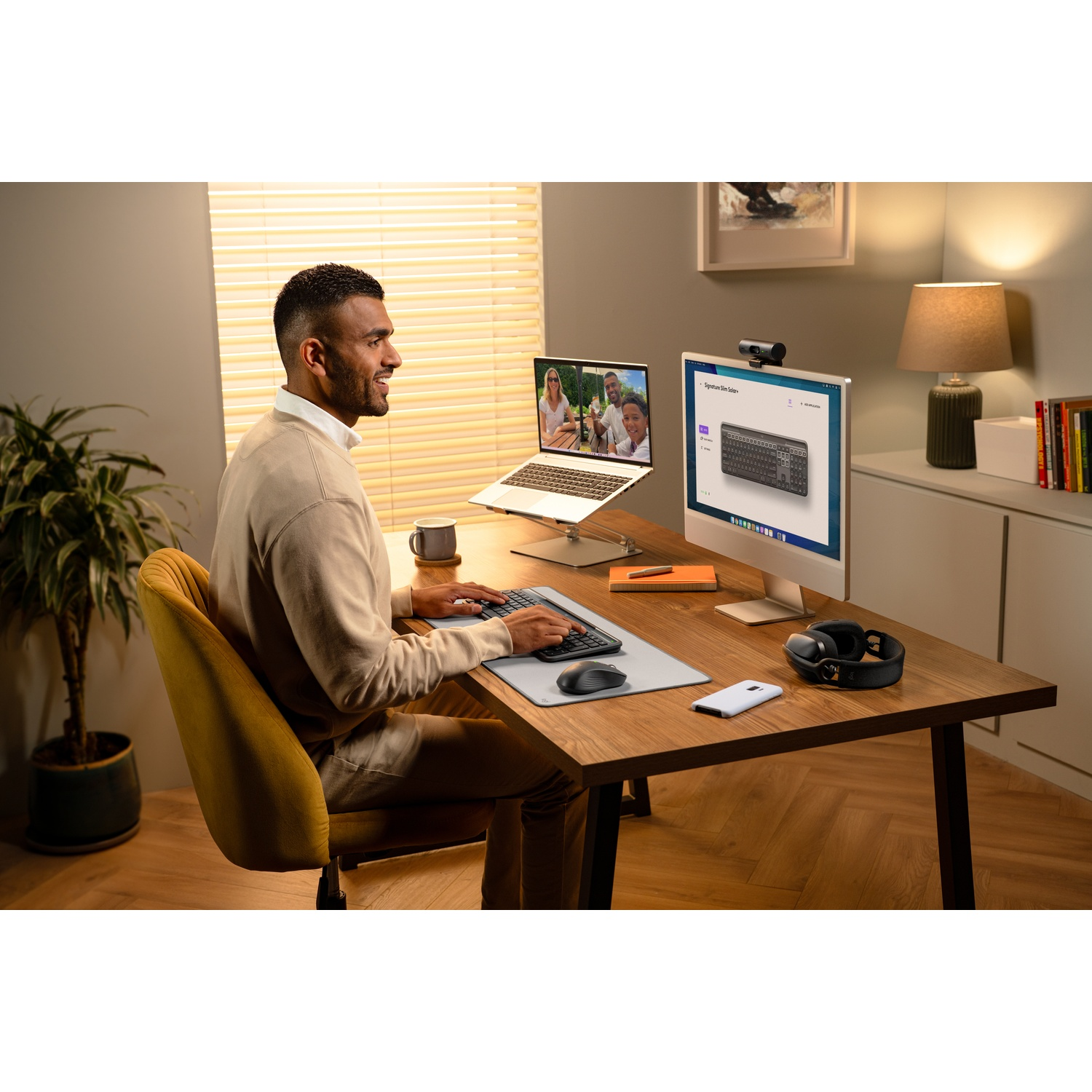 Un uomo è seduto a una scrivania con un computer, che digita. Un laptop è aperto. Una lampada e una pianta sono vicine. Una finestra con persiane è visibile.