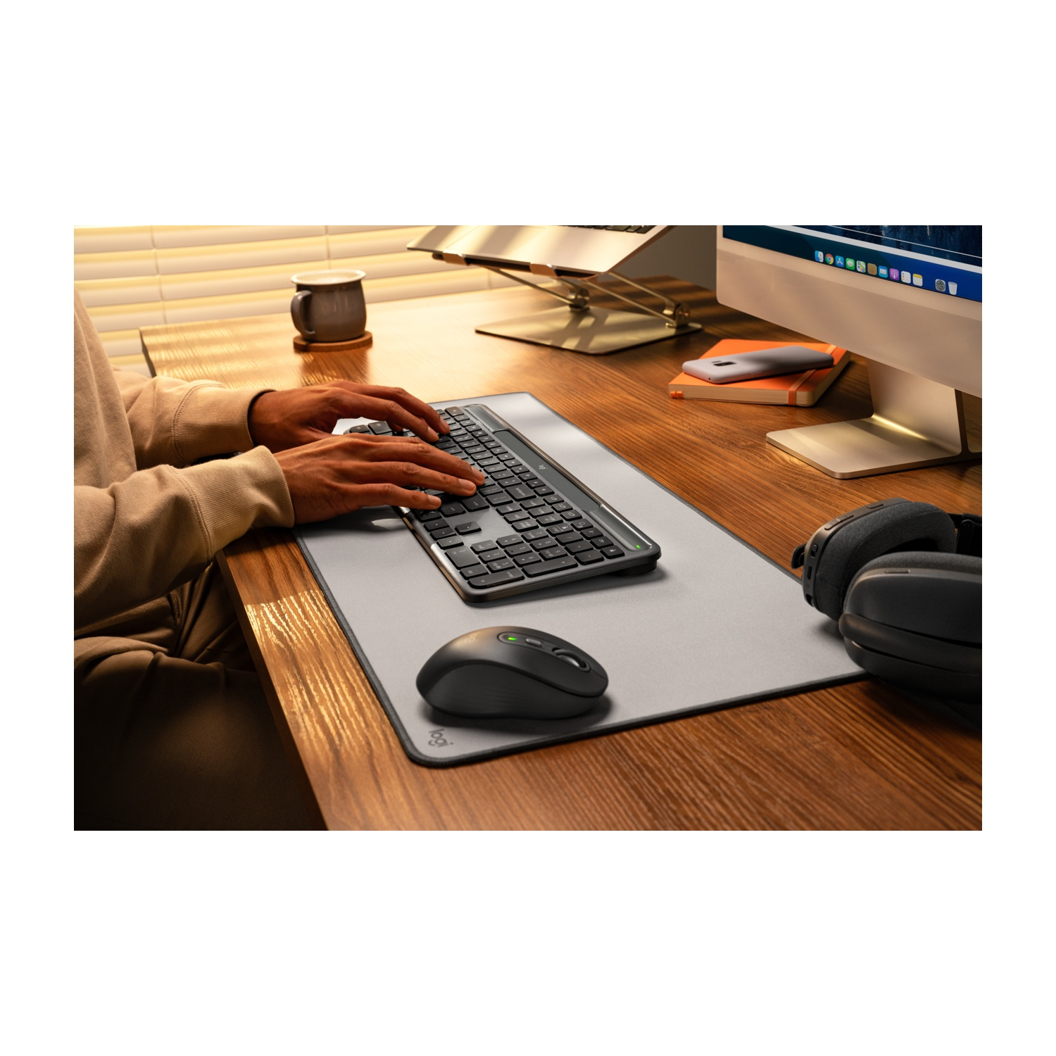 Una persona sta digitando su una tastiera a una scrivania di legno con un mouse, cuffie e un monitor. Sono presenti anche una tazza di caffè e un telefono.
