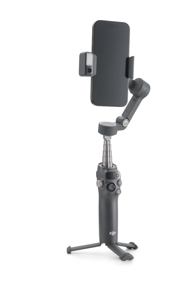 Szary telefon jest zamontowany na stabilizatorze gimbal z dołączonym małym światłem. Jest na białym tle.