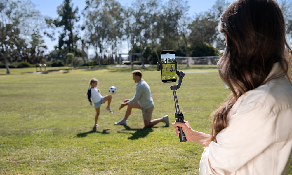 Kobieta filmuje mecz piłki nożnej telefonem na gimbalu. Dziewczyna kopie piłkę, a mężczyzna klęczy na trawie.