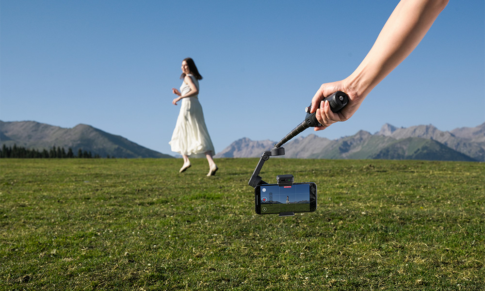 Osoba biegnie przez zielone pole. Ręka trzyma telefon na gimbalu, filmując osobę. Góry i niebo w tle.