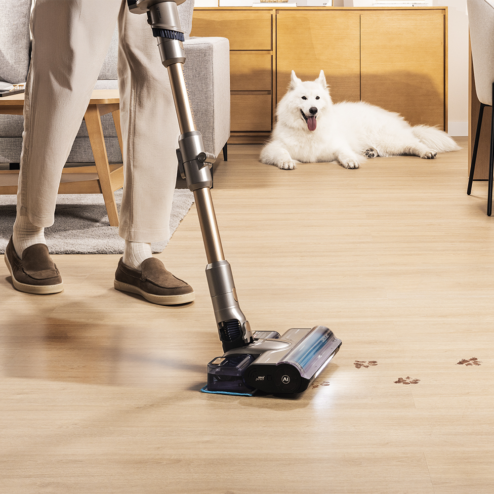 Una persona aspira un piso de madera con una aspiradora inalámbrica. Un perro blanco está en el fondo. Huellas de patas en el suelo.
