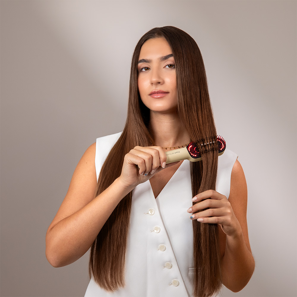 Una mujer con cabello largo castaño sostiene un cepillo para el cabello. Viste un chaleco blanco. El cepillo es beige con cerdas rojas. El fondo es gris.