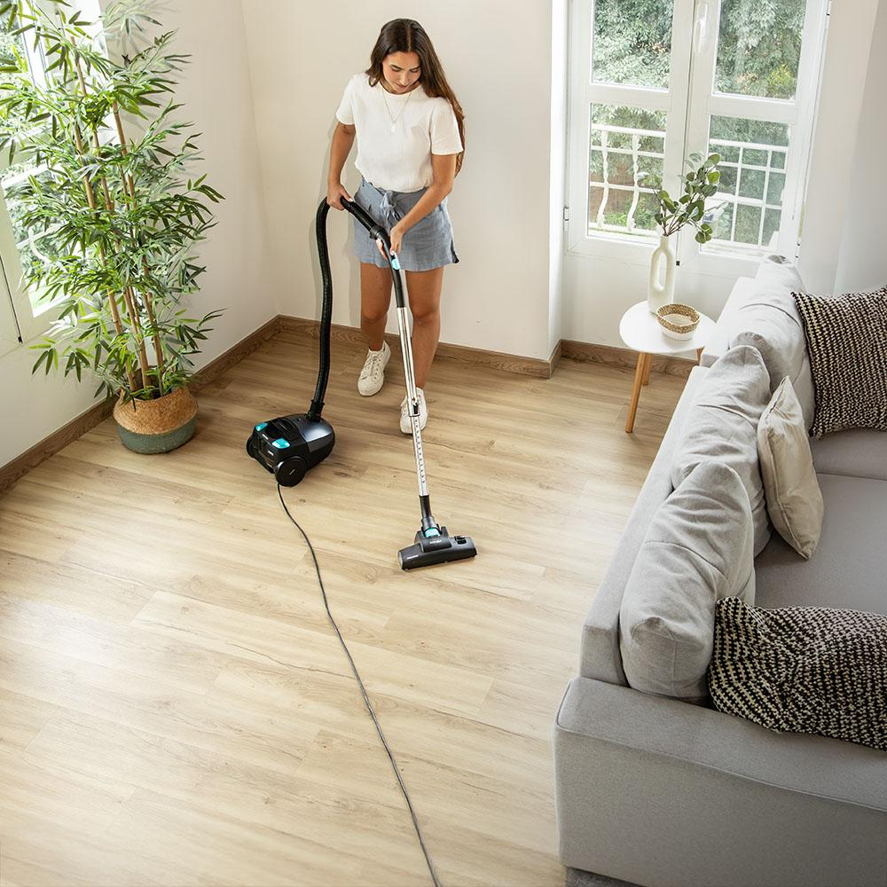 Una mujer aspira una sala de estar. Lleva camisa blanca y pantalones cortos. Un sofá, una planta y una ventana están en la habitación.