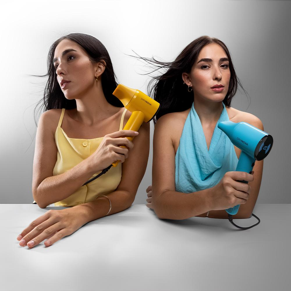 Dos mujeres sostienen cada una un secador de pelo. Una tiene amarillo y la otra azul. Están sentadas en una mesa blanca, con fondo gris.