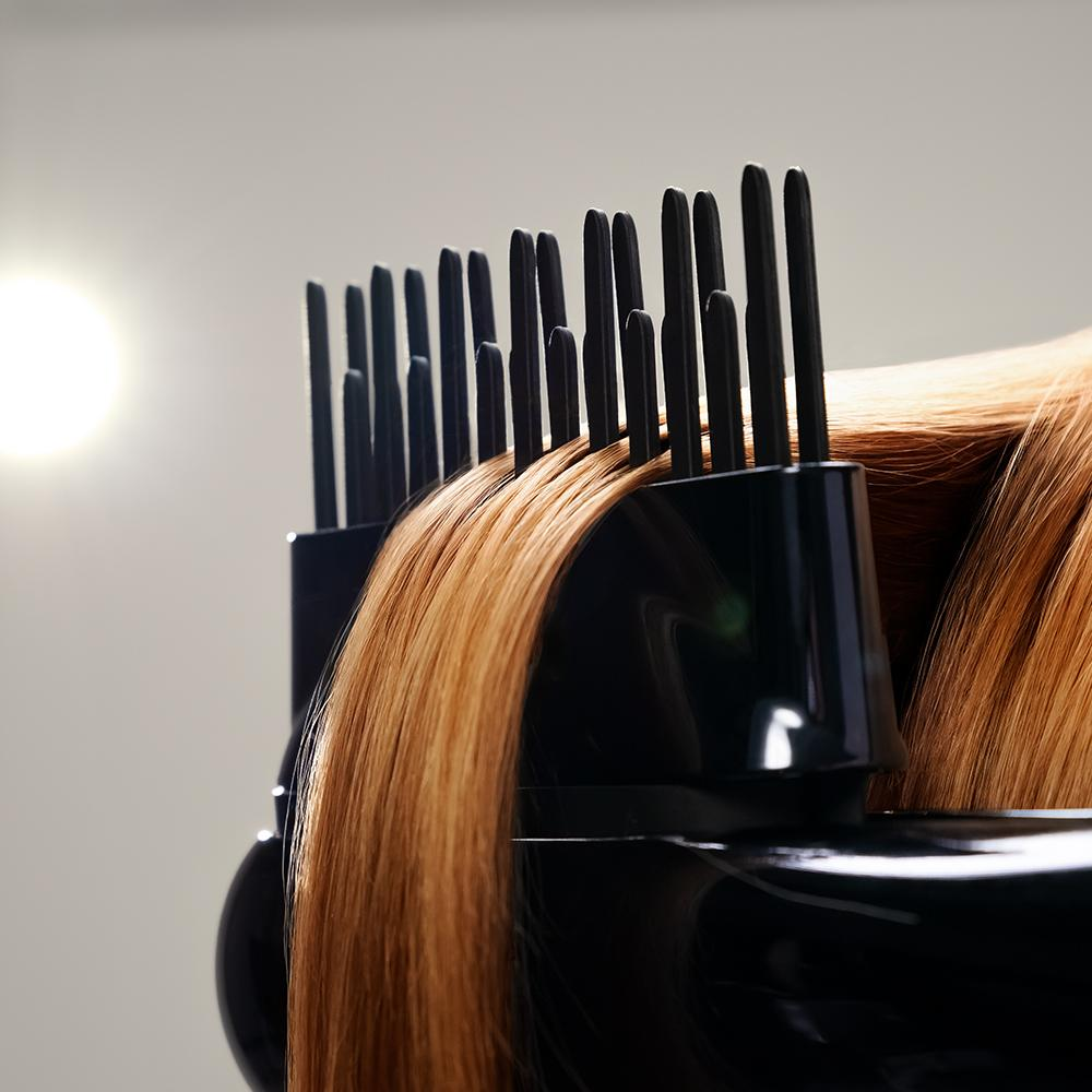 Primer plano de cabello peinándose con una herramienta negra en forma de peine. El cabello es largo, liso y castaño claro. Una luz brillante en el fondo.