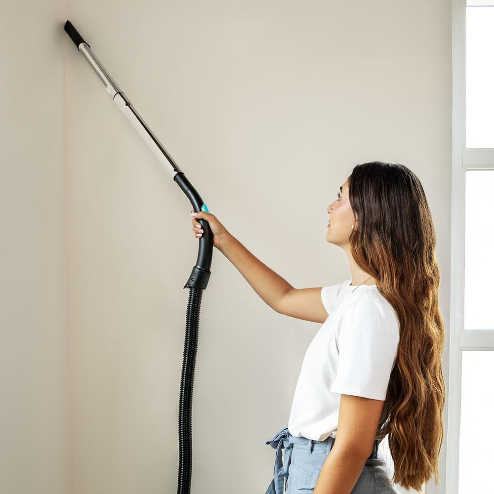 Una mujer aspira una pared con un accesorio largo de aspiradora. Viste camisa blanca y pantalones azules. Cabello largo castaño. Pared blanca.