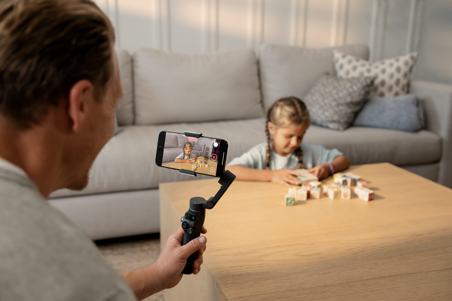Un hombre filma a una niña jugando con bloques en una mesa. La niña está en el fondo, el teléfono en primer plano.