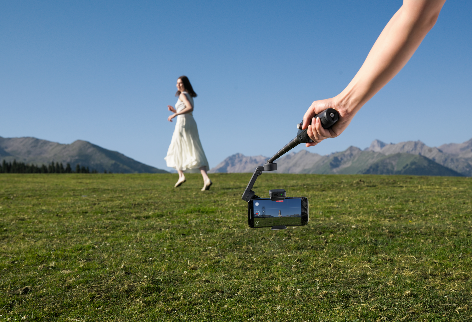 Una mujer corre en un campo. Un teléfono la graba con un cardán sostenido por una mano. Montañas y cielo azul al fondo.