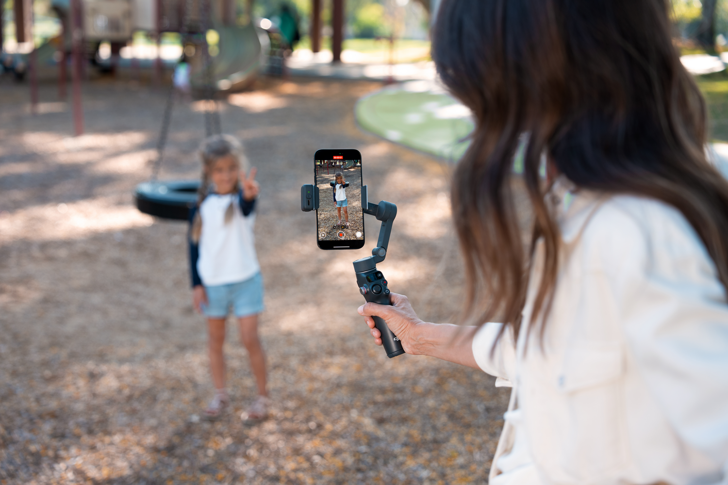 Una persona sostiene un teléfono grabando a una niña en un parque infantil. La niña en el fondo hace la señal de la victoria.