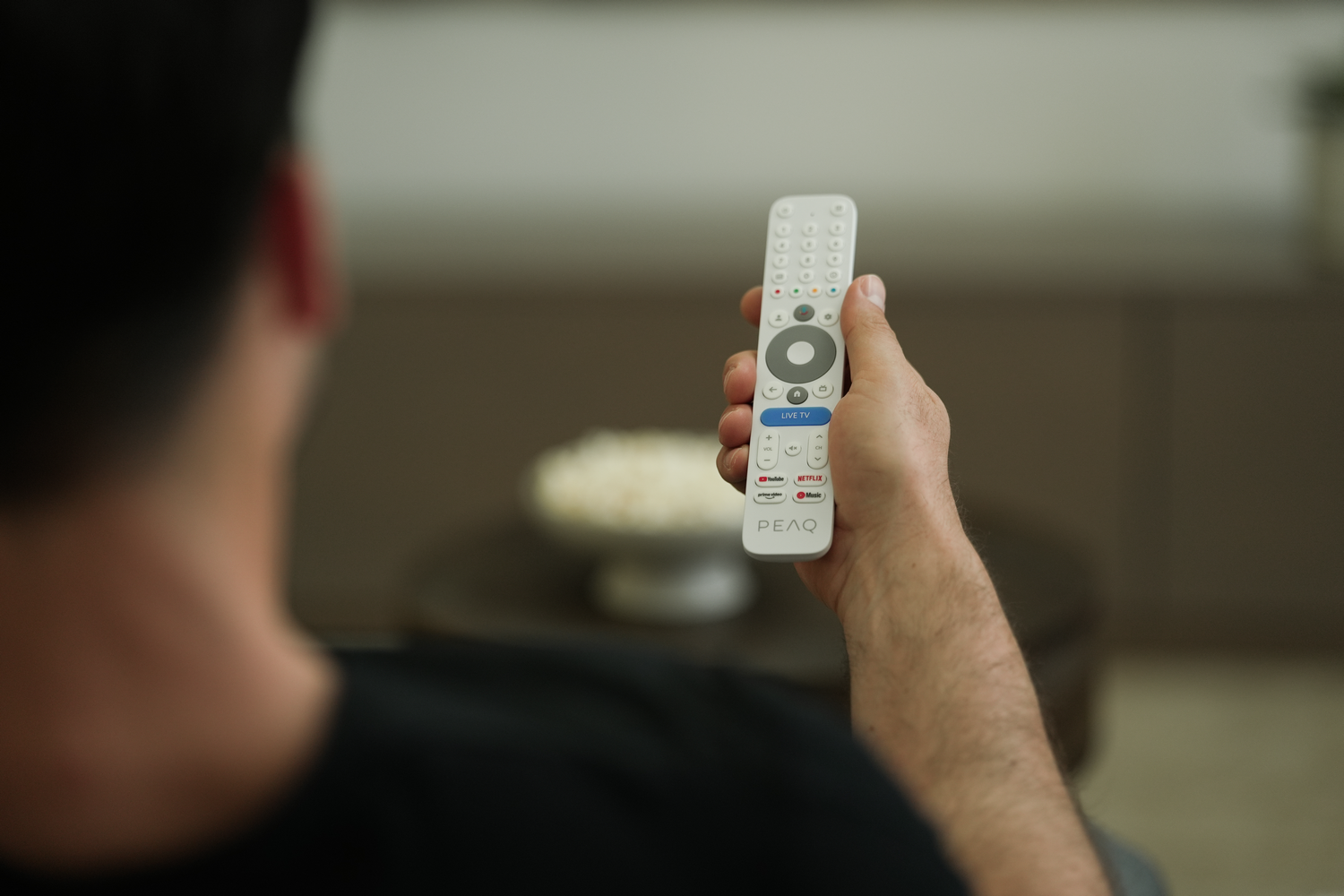Una persona sostiene un mando a distancia blanco. Un cuenco de palomitas de maíz está en el fondo. El mando tiene botones como Netflix, YouTube.