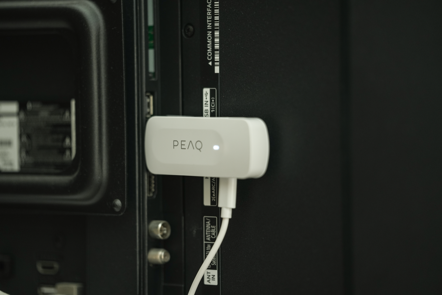 Un dispositivo PEAQ blanco está conectado a un televisor negro. El dispositivo tiene un cable USB y está etiquetado PEAQ.