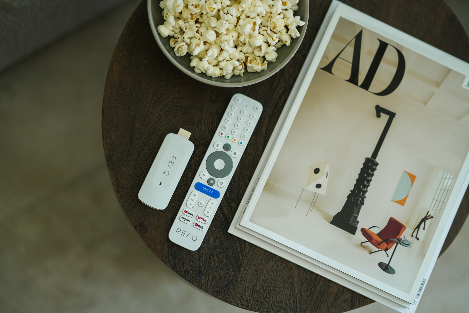 Una mesa de madera con palomitas de maíz, un mando a distancia, un dispositivo de streaming y una revista. La revista está abierta a una página con arte.