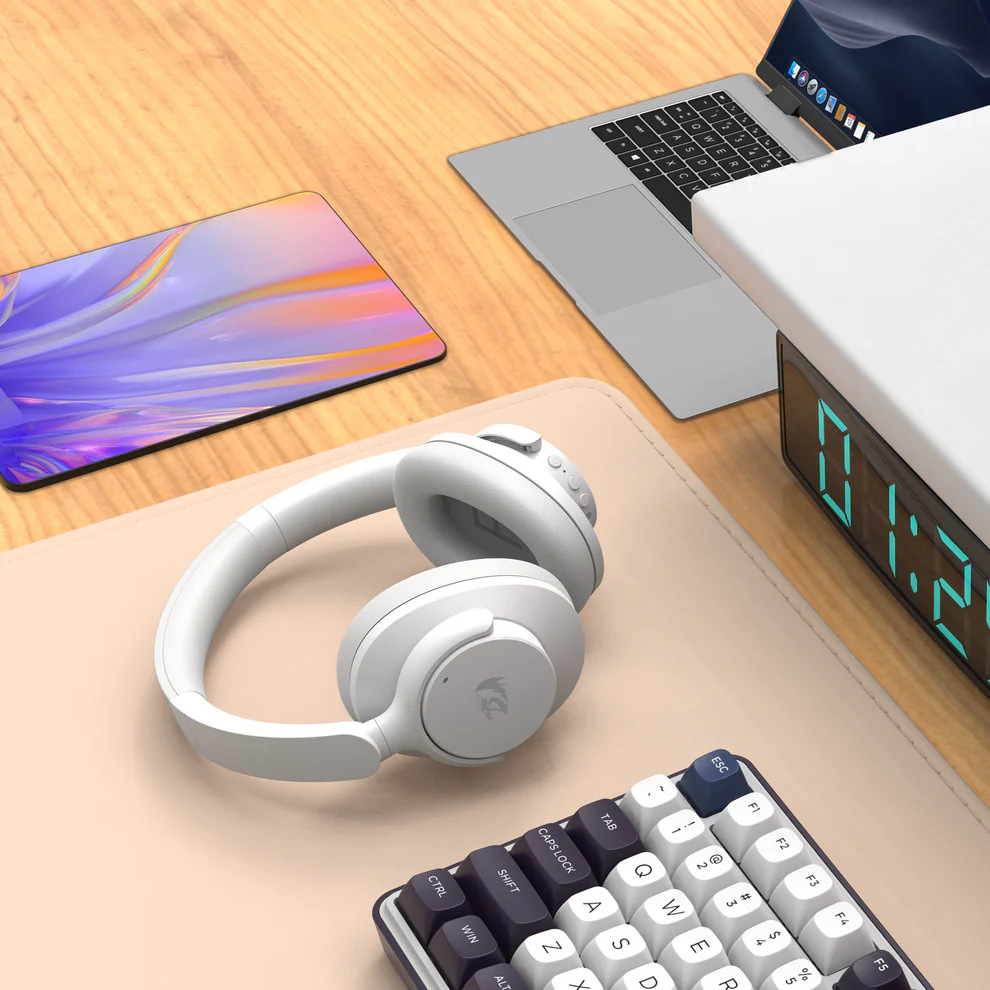 Białe słuchawki, klawiatura i laptop na biurku. Obecny jest również zegar cyfrowy i tablet.