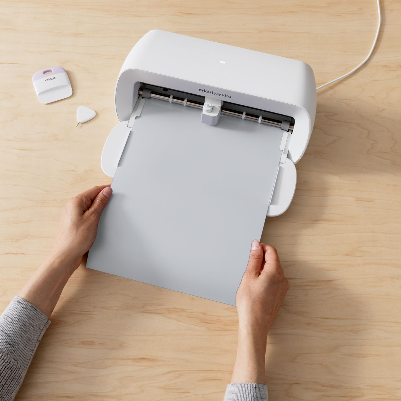 Hände halten ein graues Blatt vor einer weißen Cricut-Maschine. Werkzeuge liegen auf einer Holzoberfläche.