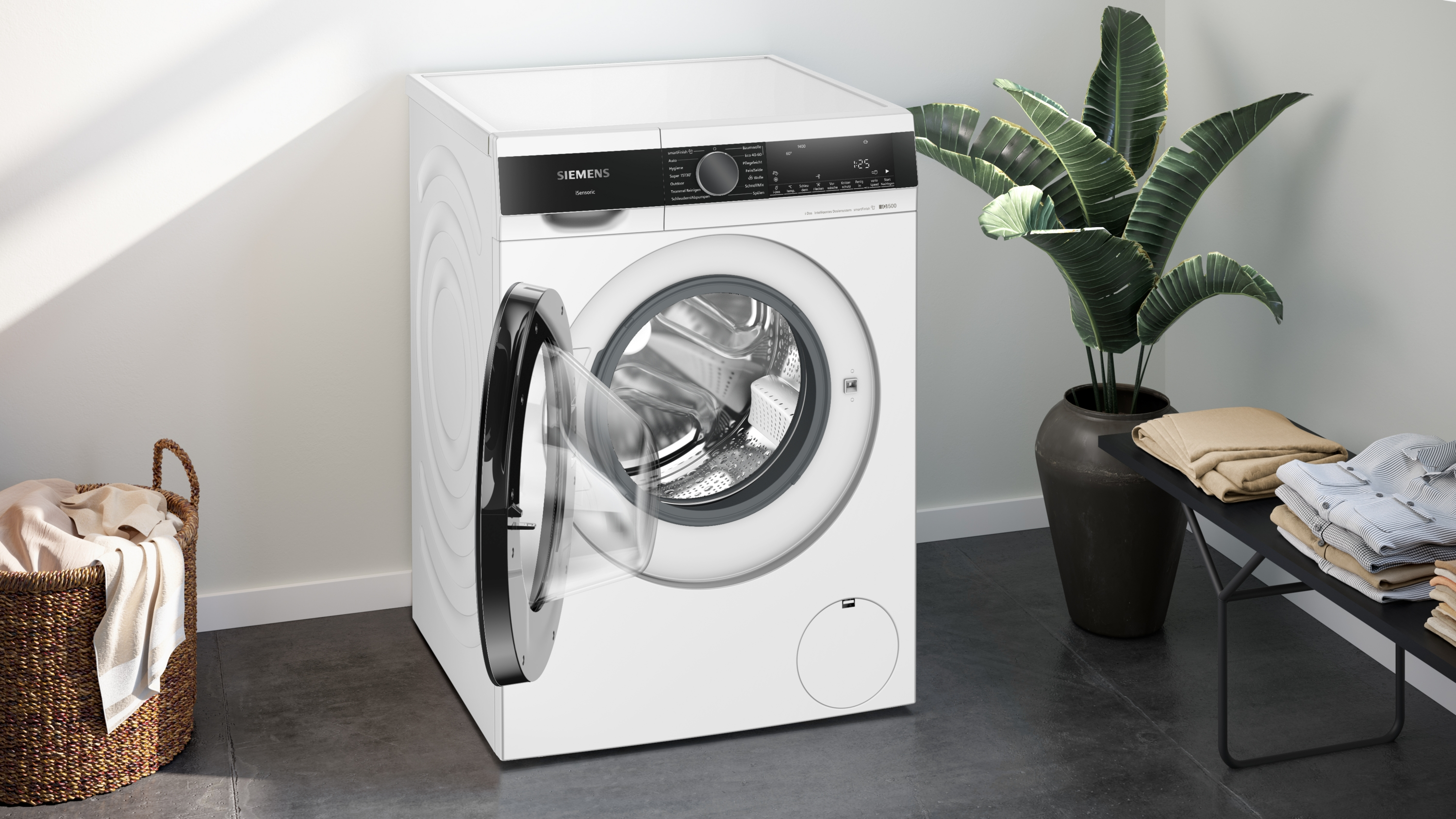 Eine weiße Waschmaschine mit offener Tür. An den Seiten befinden sich eine Pflanze und ein Korb mit Kleidung.