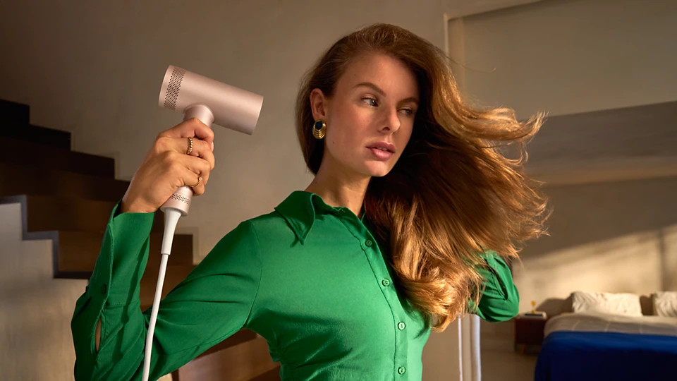 Kobieta w zielonej koszuli używa suszarki do włosów na swoich brązowych włosach. Sypialnia w tle z łóżkiem i szafką nocną.