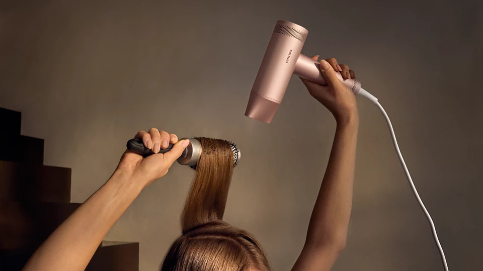 Osoba używa suszarki do włosów i okrągłej szczotki. Suszarka jest w kolorze różowego złota. Tło to ściana i schody.