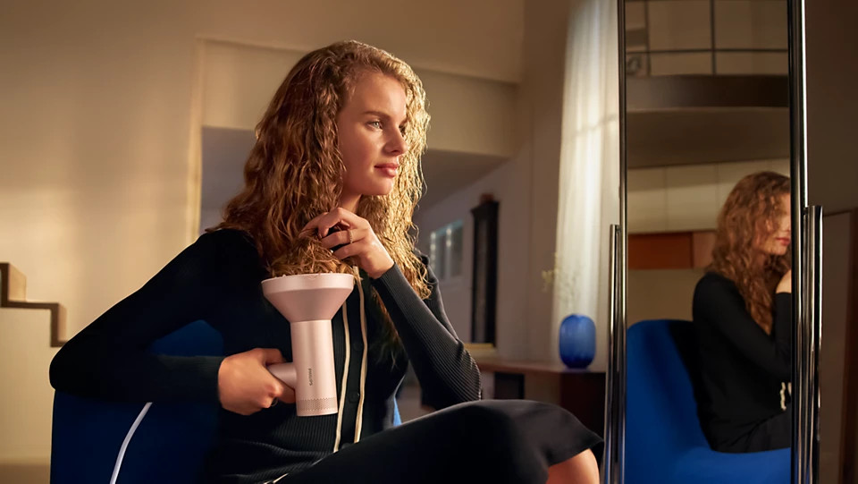 Kobieta z kręconymi włosami trzyma suszarkę, patrząc na swoje odbicie w lustrze. Siedzi na niebieskim krześle.