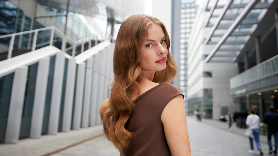 Kobieta z falowanymi włosami w brązowej sukience, spogląda przez ramię. Budynki i piesi w tle.